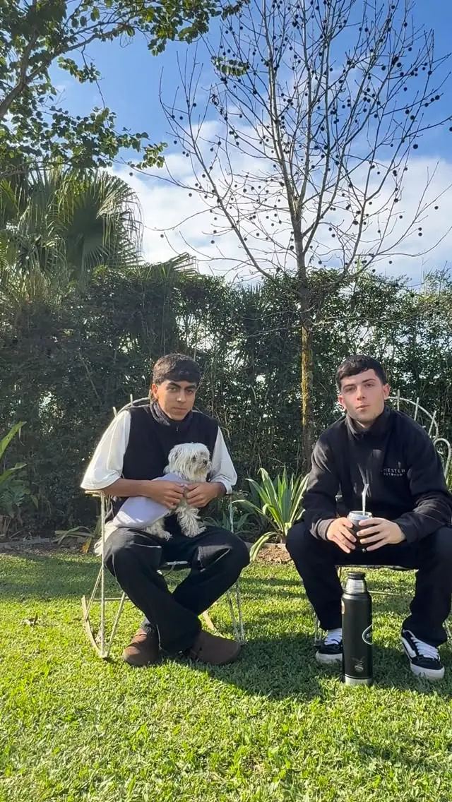 Los dos chicos de Chester Pet Shop sentados en un jardín: uno con un perrito maltés en brazos y el otro tomando mate